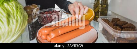 Vista dal taglio corto della donna che prende la salsiccia dal frigorifero con il cibo su sfondo bianco, scatto panoramico, immagine stock Foto Stock