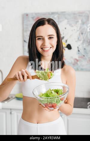 Bella sportswoman sorridente alla macchina fotografica mentre tiene la ciotola con insalata fresca in cucina Foto Stock