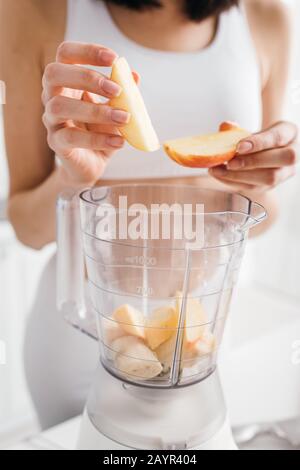 Vista corta di giovane sportswoman mettere la frutta nel frullatore sul tavolo da cucina Foto Stock