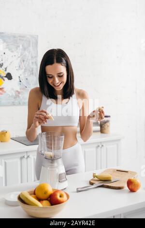 Bella sportswoman sorridente mettere banana nel frullatore mentre si prepara frullato Foto Stock