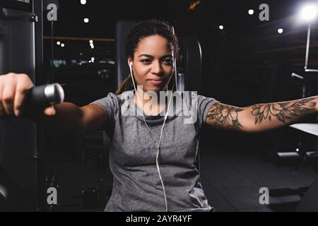 fuoco selettivo di felice e tatuato donna afroamericana ascoltare musica in palestra Foto Stock