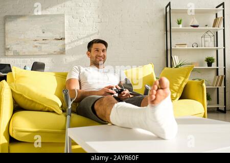 Kyiv, UCRAINA - 21 GENNAIO 2020: Bel uomo sorridente con gamba rotta sul tavolino da caffè giocando a video gioco in soggiorno Foto Stock