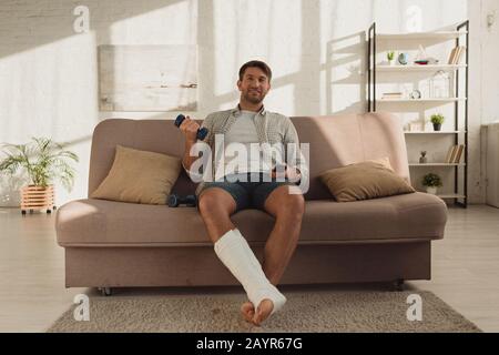 Uomo sorridente con gamba rotta guardando la tv e tenendo dumbbell sul divano a casa Foto Stock