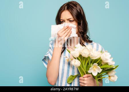 donna malata che asciuga il naso con tovagliolo di carta mentre tiene tulipani bianchi isolati su blu Foto Stock