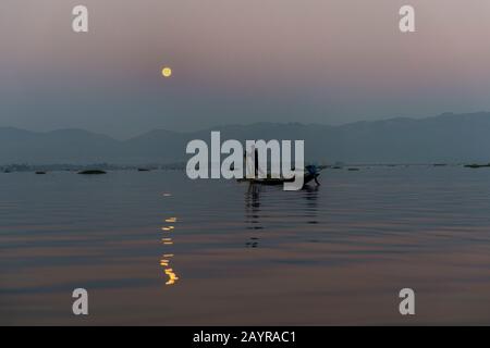Prima dell'alba sotto una luna piena un pescatore di canottaggio della gamba nella sua barca sta tirando nella sua rete di pesca ha regolato il giorno prima sul lago di Inle in Myanmar. Foto Stock