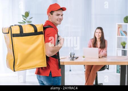 Fuoco selettivo di uomo di consegna con termo zaino sorridente alla macchina fotografica con businesswoman al tavolo in ufficio Foto Stock