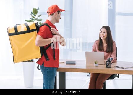 Attraente donna d'affari sorridente alla consegna uomo con termo zaino mentre lavora al tavolo Foto Stock