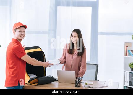 Consegna uomo con borsa termica che dà contenitore di cibo a donna d'affari e sorridente alla macchina fotografica in ufficio Foto Stock