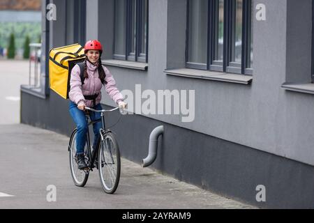 Corriere sorridente con termo zaino in bicicletta vicino all'edificio Foto Stock