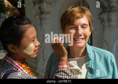 Una giovane donna sta mettendo la pasta fatta dall'albero di Thanaka su una faccia di un turista alla Pagoda di Kutodaw sulla collina di Mandalay, Mandalay, Myanmar. Foto Stock