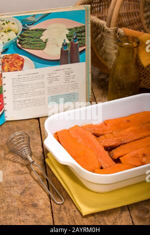 Patate dolci candite al forno in una teglia rettangolare su un piano rustico in legno con un vecchio libro di cucina, whisky antico e cesto sullo sfondo Foto Stock