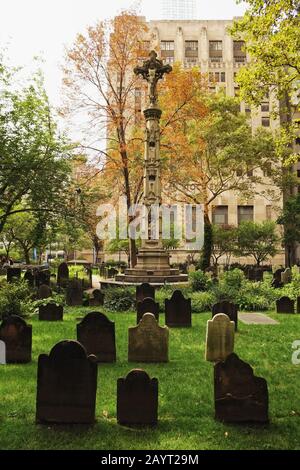 Trinity Churchyard Manhattan NYC le lapidi in fila intorno a una colonna di scultura sormontata da Cristo sulla croce, erba verde e alberi Foto Stock