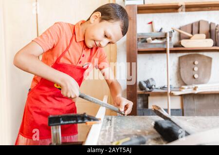 Piccolo geniale ragazzo di talento lavora con il legno in un laboratorio di Falegnameria. Il concetto di apprendimento, hobby e lavoro manuale manuale per i bambini Foto Stock