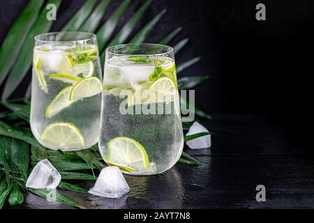 Mojito in un grande vetro trasparente con foglie di lime e menta e cubetti di ghiaccio. Sullo sfondo sono rami di palma. Su uno sfondo di legno nero. Copia Foto Stock