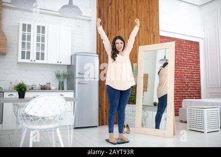 Giovane donna in una blusa beige che si erge su una pesatrice e si sente grande Foto Stock