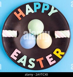 Buon testo pasquale con uova colorate e marshmallows disposti su piastra nera - vista dall'alto e formato quadrato Foto Stock