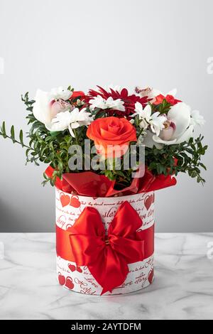Bouquet di fiori colorati in una scatola con fiocco rosso a nastro su sfondo grigio. Regalo, biglietto d'auguri Foto Stock