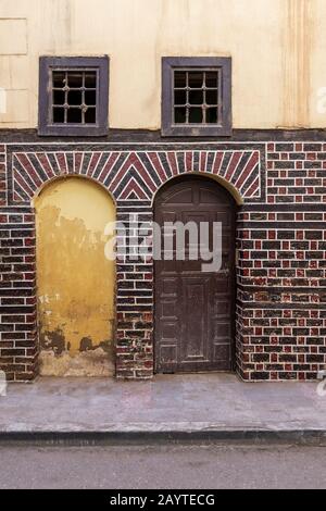 Porta in pietra bloccata, porta a volta decorata in legno, e due piccole finestre con barre in ferro arrugginito su mattoni neri e rossi muro su strada lastricata Foto Stock
