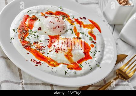 Uova turche con yogurt greco e salsa al burro di pepe aleppo, cilbir, servite su piatti bianchi con posate dorate su un tavolo di legno bianco, orizzontale Foto Stock