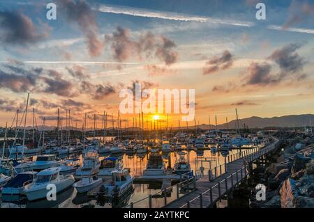 Tramonto epico sulla città di Coffs Harbour sulla costa australiana con yacht e barche in una baia e montagne in lontananza, Foto Stock