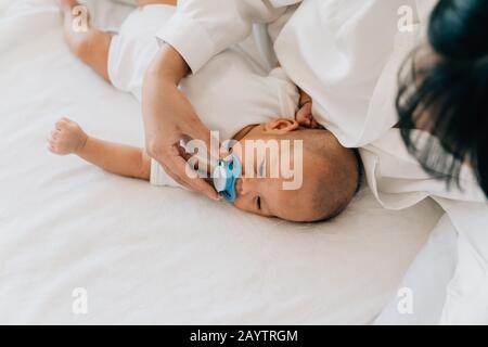 Dall'alto della mamma premurosa che dà il succhietto al neonato asiatico piccolo che mette il succhietto nella calma bocca del neonato sdraiata sul letto della casa Foto Stock