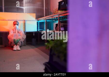 Ritratto a tutta lunghezza dello scienziato femminile che indossa un equipaggiamento protettivo completo seduto sul pavimento in bio-laboratorio illuminato da luce UV, copia spazio Foto Stock