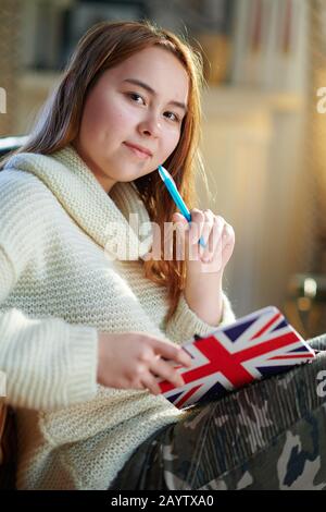 Ragazza adolescente moderna con capelli rossi in maglione bianco nella casa moderna nella soleggiata giornata invernale scrivere nei colori del notebook della bandiera britannica. Foto Stock