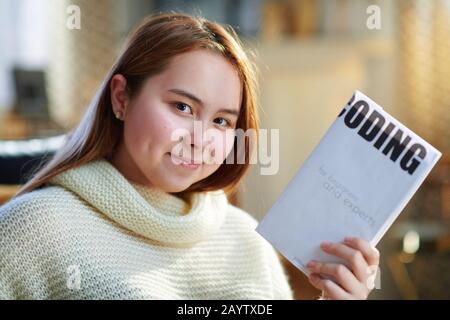 sorridente ragazza moderna adolescente con capelli rossi in maglione bianco nella casa moderna nella soleggiata giornata invernale mostrando libro didattico di codifica. Foto Stock