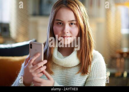 Ritratto di giovane ragazza sorridente moderna con capelli rossi in maglione bianco nel moderno soggiorno nella soleggiata giornata invernale utilizzando smartphone per onlin sociale Foto Stock