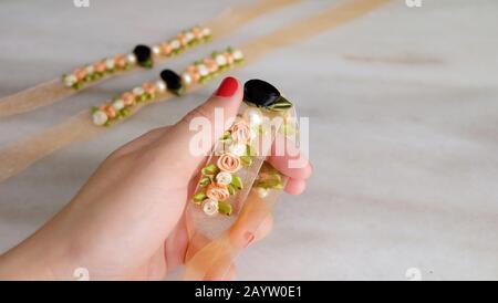 Una mano che tiene un corsalvia di fiore fatto con i fiori del nastro e le perle false, con due corsates più simili nello sfondo. Foto Stock