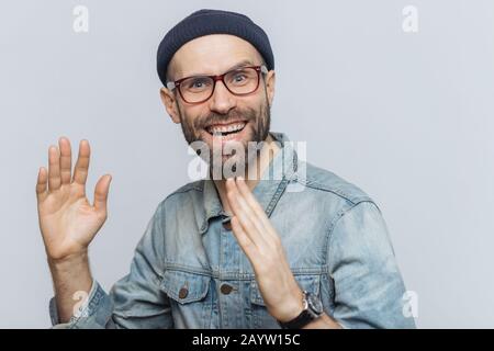 Ritratto di felice uomo di mezza età ha divertimento indoor, alza le mani, ha il piacere di espressione, indossa gli occhiali, cappello e denim shirt, isolato su bianco backgr Foto Stock