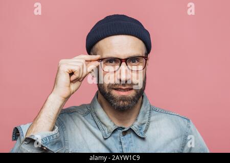 Ritratto di piacevole guardando soddisfatto uomo barbuto con un aspetto gradevole guarda con fiducia attraverso occhiali, indossa abbigliamento di moda, pone ag Foto Stock
