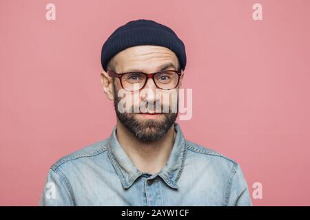 Ritratto di bello uomo barbuto guarda con lieta sorpresa espressione, indossa gli occhiali e cappello nero, isolato su rosa di sfondo per studio. Centro ag Foto Stock