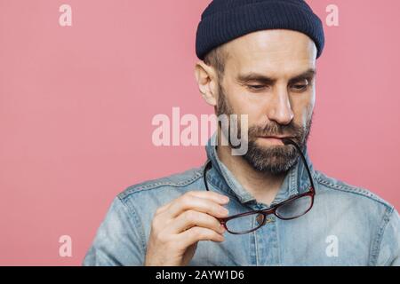 Colpo di Testa di pensieroso barbuto uomo guarda pensieroso verso il basso, mantiene gli occhiali, indossa camicia denim e hat, isolate su sfondo rosa con copia spazio per Foto Stock