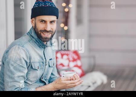 Ritratto di bel maschio sorridente giovane gode di tempo libero, tiene tazza di caffè, essendo profondo nei pensieri, ha pausa caffè durante la giornata calda brillante, focu Foto Stock