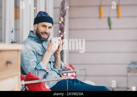 Positivo felice attraente modello maschile tiene una tazza di tè, ha un buon riposo al balcone, respira aria fresca e gode di sole tempo, ha sorriso piacevole su fac Foto Stock