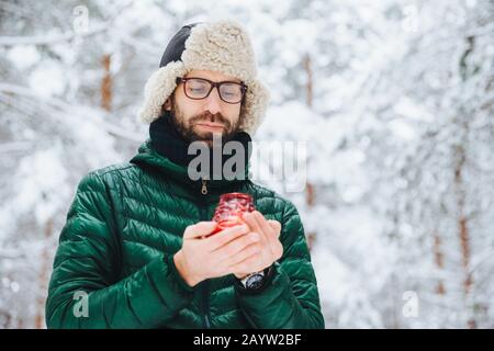 Attraente maschio gravi indossa caldo abbigliamento invernale, mantiene la candela, si erge contro l'inverno sfondo di foresta, gode di aria fresca, essendo molto fiducioso. Fash Foto Stock