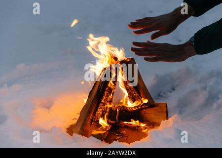 Maschio irriconoscibile scalda le mani sul fuoco nella foresta durante il freddo inverno, prova a caldo se stesso, essendo freddo trascorrere del tempo sulla neve fredda e gelido meteo. Bu Foto Stock