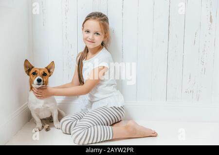 Bella femmina piccolo bambino gioca con il suo cane in camera bianca, sedersi sul pavimento, hanno buon rapporto, coccole animale preferito. Little Schoolgirl ama anima Foto Stock