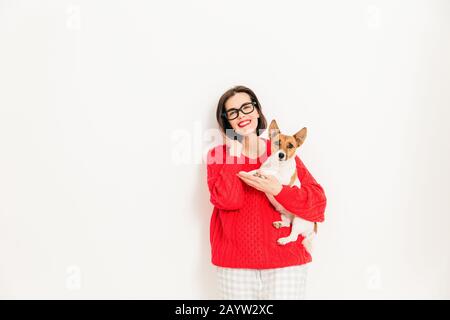 Giovani caucasici modello femminile con felice espressione, indossa gli occhiali e maglione rosso, mantiene la sua preferita jack russell terrier cane, isolato su bianco Foto Stock