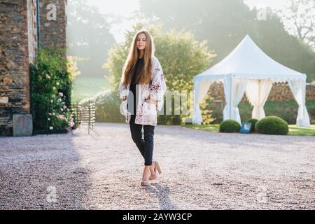 Ritratto orizzontale all'aperto di donna piacevole osservare alla moda sulle scarpe alte heeled, guarda penosamente giù, posa alla macchina fotografica, si levano in piedi contro meraviglioso Foto Stock