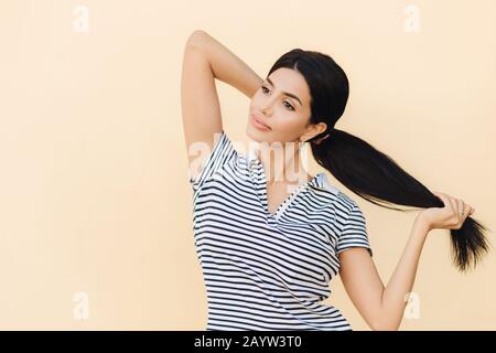 La femmina dai capelli scuri e penosa rende la coda di pony, ha labbra piene e aspetto piacevole, indossa abiti casual, si leva sullo sfondo dello studio, focalizzato in Foto Stock