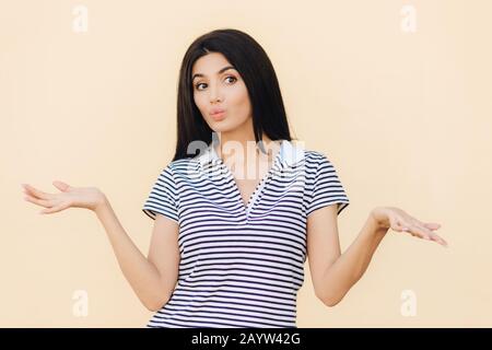 Bella femmina ha espressione senza clubs, spalline, tiene le labbra rotonde, guarda esitante da parte, vestito in abiti casual, isolato sopra lo studio ba Foto Stock