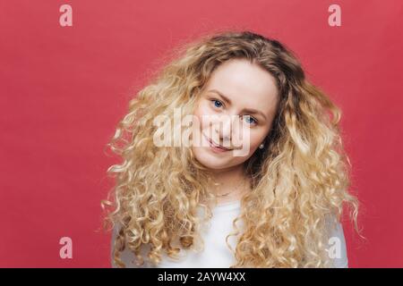 Attraente femmina con capelli curly bush e occhi blu, guarda positivamente alla fotocamera, si sente soddisfatto come incontro con gli amici, isolato su ba rosa brillante Foto Stock