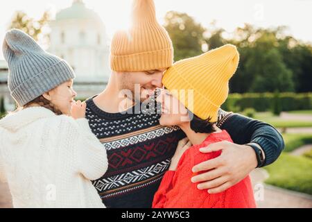 Attraente uomo indossa giallo caldo hat, abbraccia la moglie e la figlia, guarda a loro con grande amore. Adorabile bambina si sente sostegno da parte di genitori, Foto Stock