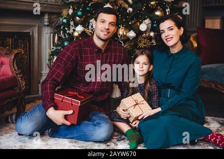 Padre, madre e figlia godersi vacanze insieme, sorriso felicemente come sedersi in soggiorno vicino decorate Anno nuovo albero, tenere presente confezionate in scatole, Foto Stock