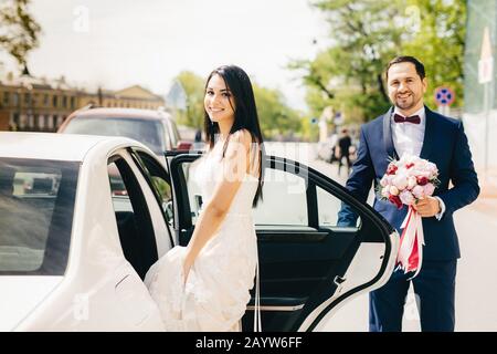 Ritratto di coppia newlywed sedersi in auto dopo la cerimonia, hanno felice espressione. Sorridente sposa femminile in abito bianco si siede in auto bianca, andando ad avere cru Foto Stock