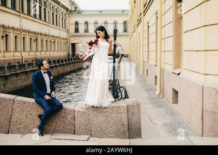 Bel sposo si siede sul ponte guarda la sua bella sposa che si alza, dice complimenti a lei, godere insieme, avere momenti indimenticabili Foto Stock