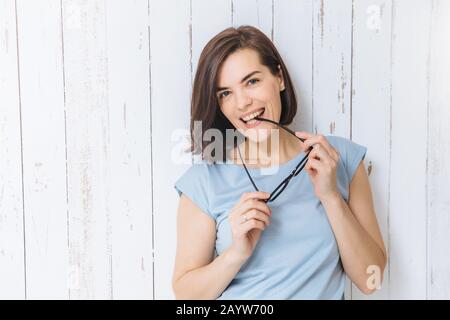 Vita ritratto di blue eyed brunette femmina si toglie gli occhiali vestita casualmente si erge contro il bianco sullo sfondo di recinzione, essendo di buon umore. Chee Foto Stock