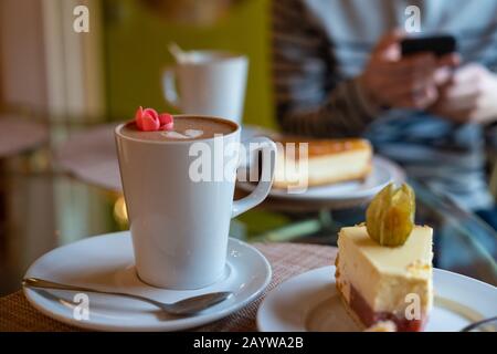 Primo piano di una tazza bianca di latte di caffè decorato con un cuore e un fiore, torta con boccone preso. Uomo che tiene lo smartphone sullo sfondo. Foto di scorta Foto Stock